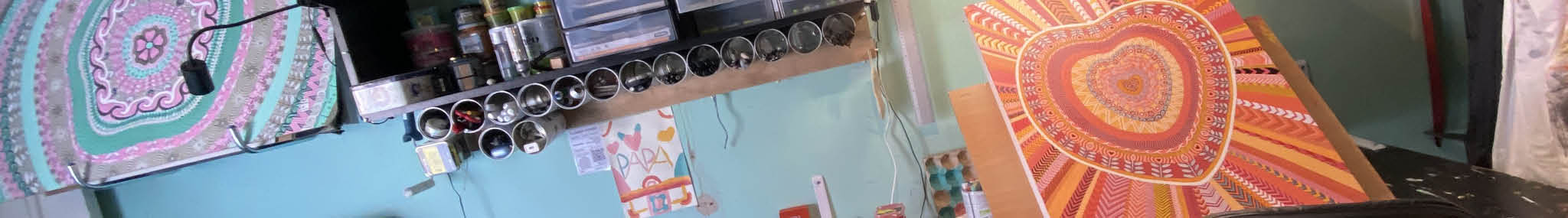 bannière artwork pour la section des tableaux originaux. photo prise dans l'atelier de l'artiste fabien trarieux où l'on aperçoit une peinture en cours, une autre terminée fixé au mur et plusieurs boites de café accrochées à des étagères pour y faire des rangements pour pinceaux, feutres, gommes...